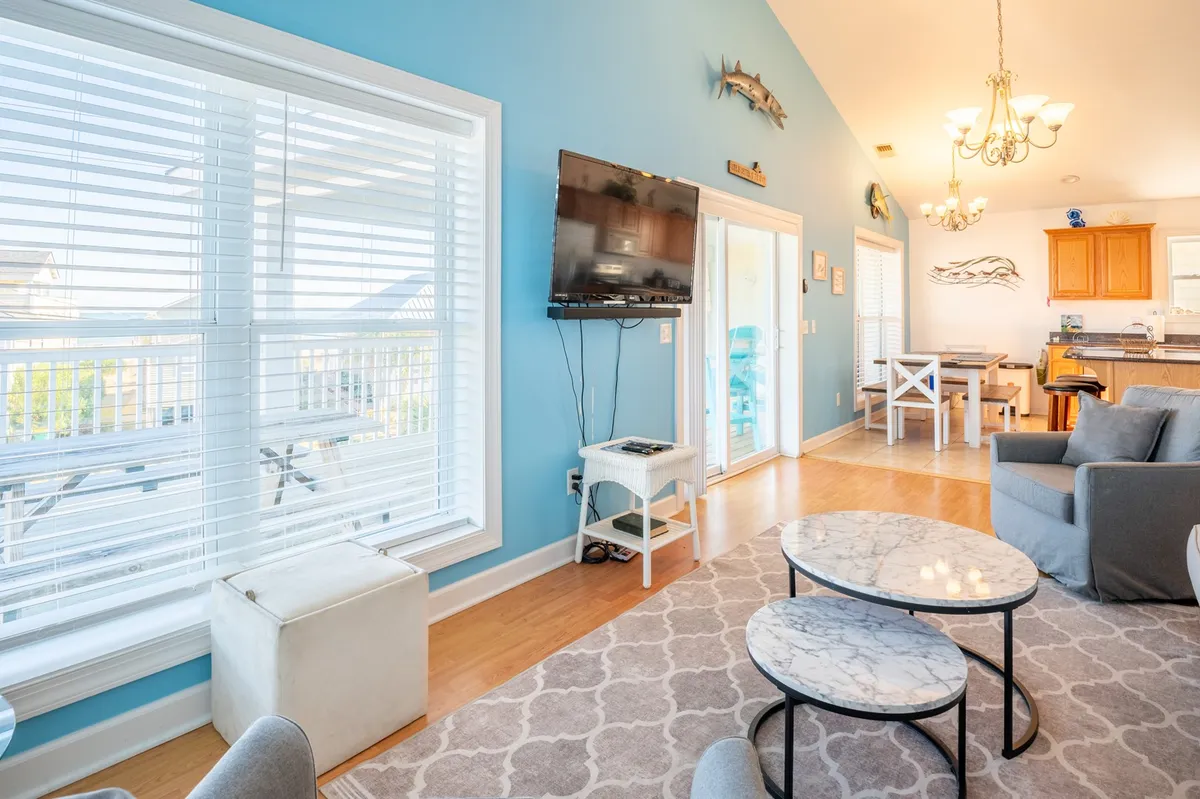 A welcoming living area with large windows, a marble coffee table, blue walls, a television, and a statement light fixture. The space feels sunny, open, and perfect for socializing or enjoying a morning coffee.