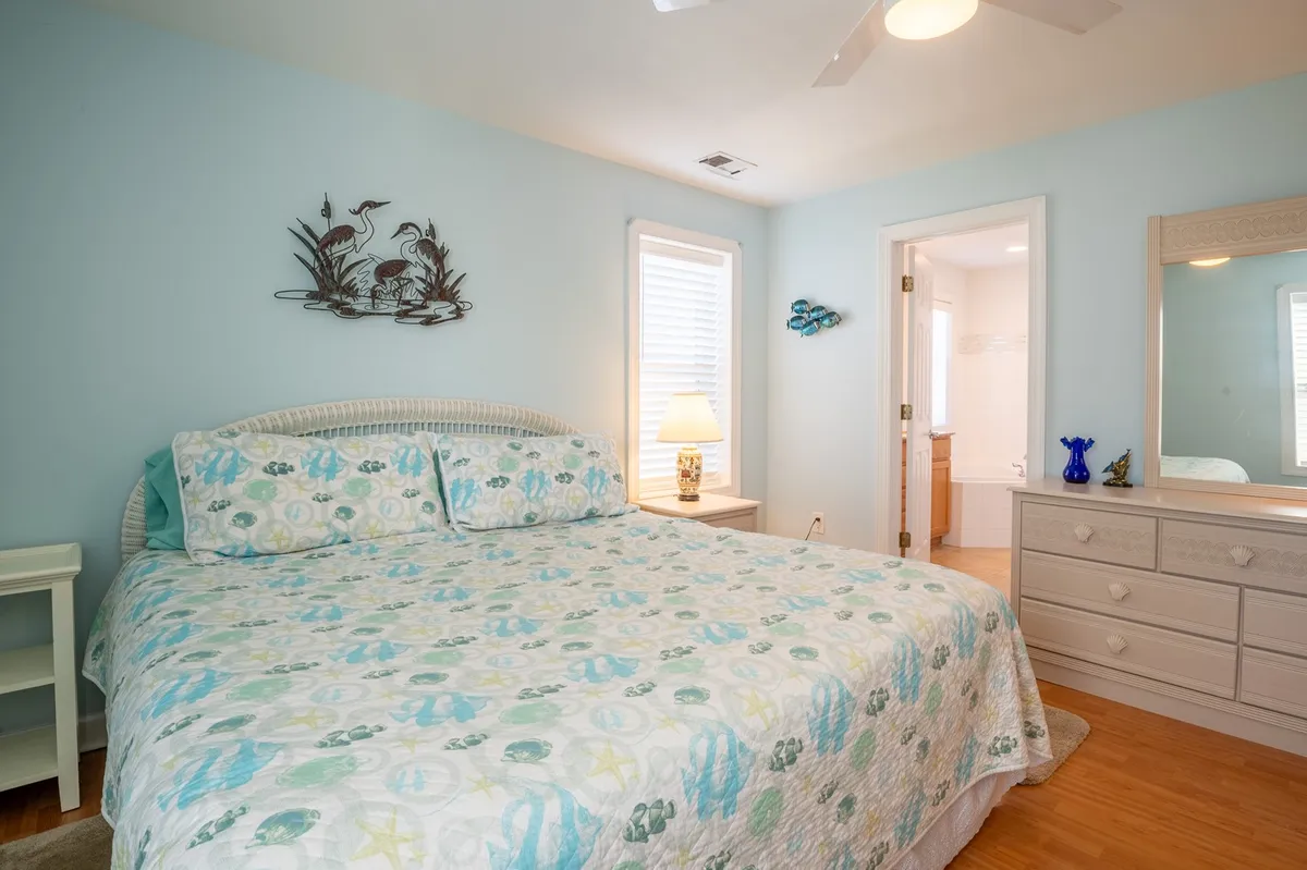 A tranquil bedroom scene featuring a bed with a colorful quilt, decorative bird wall art, and soft lamp lighting. Natural light seeps in, suggesting a restful, private escape to unwind after a day of beach exploration.