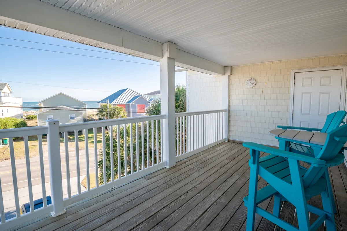A covered porch with bright blue Adirondack chairs and a white railing overlooks the ocean. The space is inviting, tranquil, and perfectly poised for enjoying a coastal breeze, reading, or sipping coffee while taking in the view. Sunlight filters onto the porch, creating an ideal spot for relaxation at any time of day.
