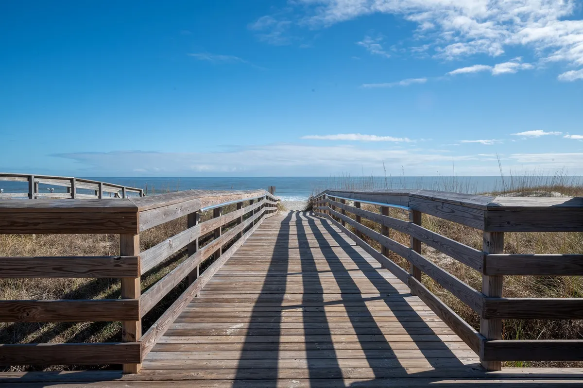 A scenic wooden walkway leads toward the expansive ocean, framed by soft clouds in the sky. The tranquil scene captures the inviting shoreline, perfect for morning strolls or family shell-collecting adventures.