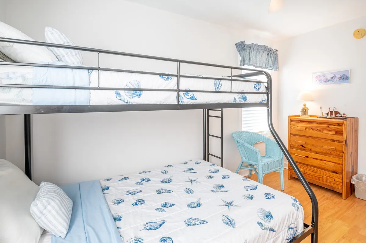 A cheerful bunk room with a twin-over-queen bunk bed dressed in blue shell-patterned bedding, a wooden dresser, and a light blue chair by the window. Natural light fills the space, making it both playful and relaxing.
