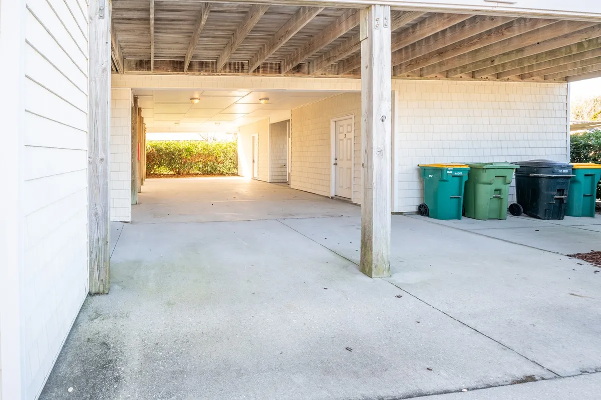 A ground-level exterior view shows an open parking pad beneath the home, featuring designated space for vehicles, support beams, and garbage bins. The area is uncluttered and practical, highlighting ease of arrival and efficient use of space for parking and storage.
