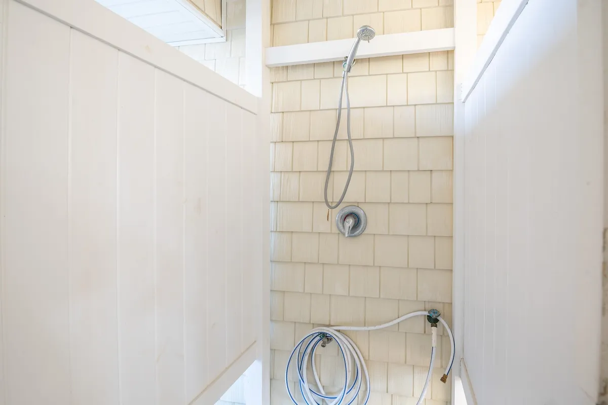 An outdoor shower fixture is mounted on a wall of white wooden panels and textured tiles. The simple, coastal design evokes post-beach rituals—rinsing off after sun-drenched adventures before heading inside. Sunlight brightens the area, underscoring its practicality and charm.