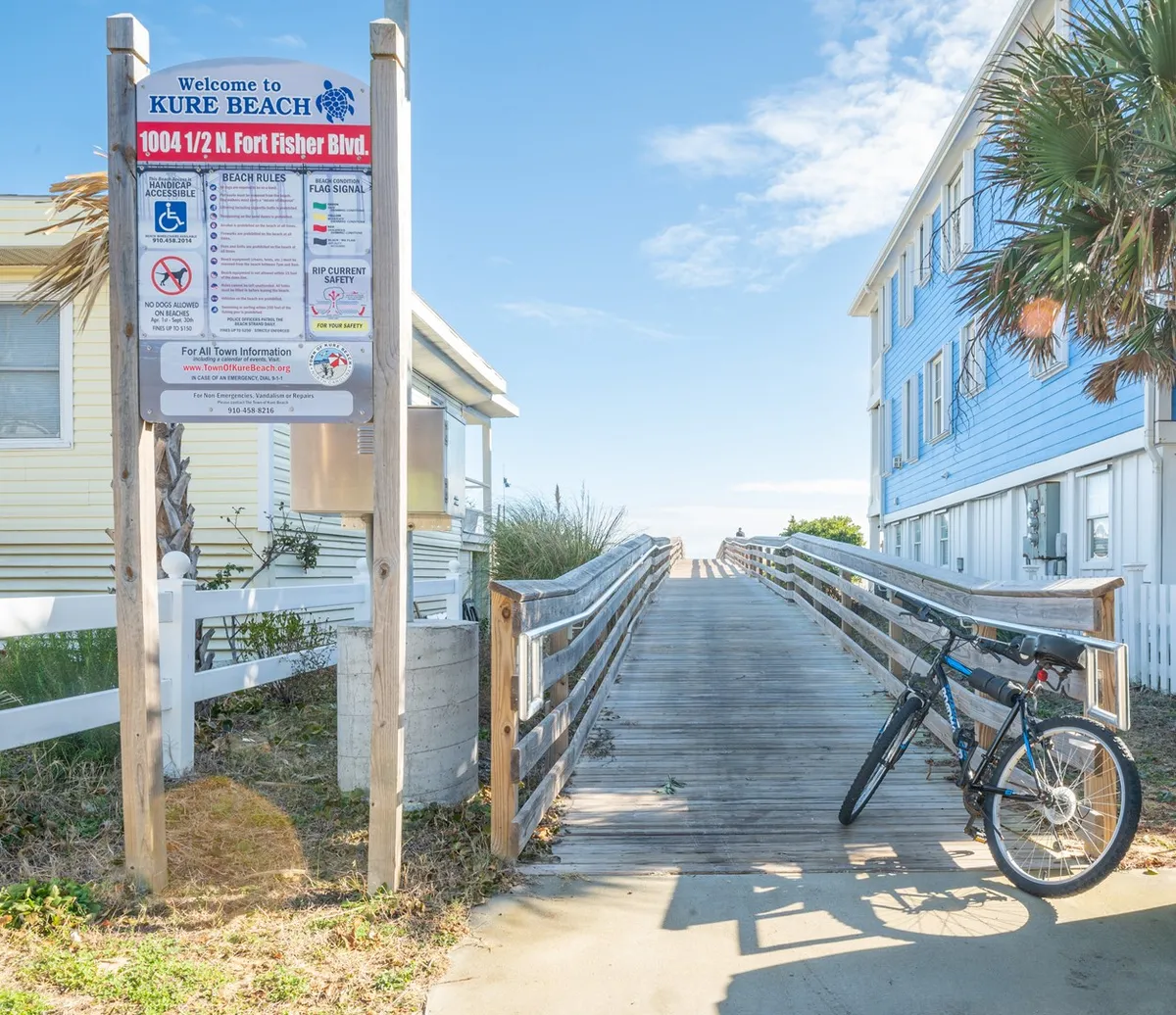 A wooden walkway bordered by a Kure Beach sign and a bicycle, setting a relaxed, welcoming mood. The boardwalk hints at casual beachside exploration, morning bike rides, and the family-friendly vibe of the area.