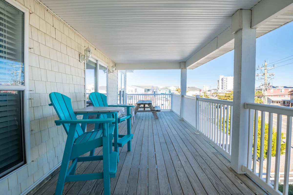 An inviting exterior scene features several blue Adirondack chairs arranged around a wooden picnic table on a spacious balcony. The area is ideal for gathering, with open railings revealing distant ocean views and plenty of space for group activities, morning coffee, or relaxed evening conversations. The tranquil mood is set by the fresh sea breeze, warm lighting, and the promise of sunsets visible from the porch.