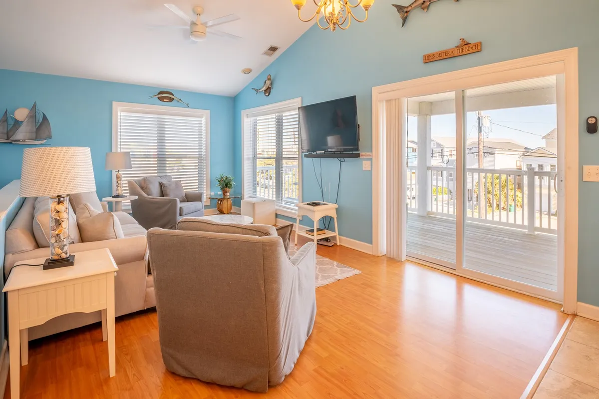 A sun-filled living room with light blue walls, a plush sofa, and a Smart TV. Large sliding glass doors open to reveal an inviting outdoor view, creating a seamless indoor-outdoor feel. The space is arranged for relaxation and conversation, filled with cheerful, relaxed energy.