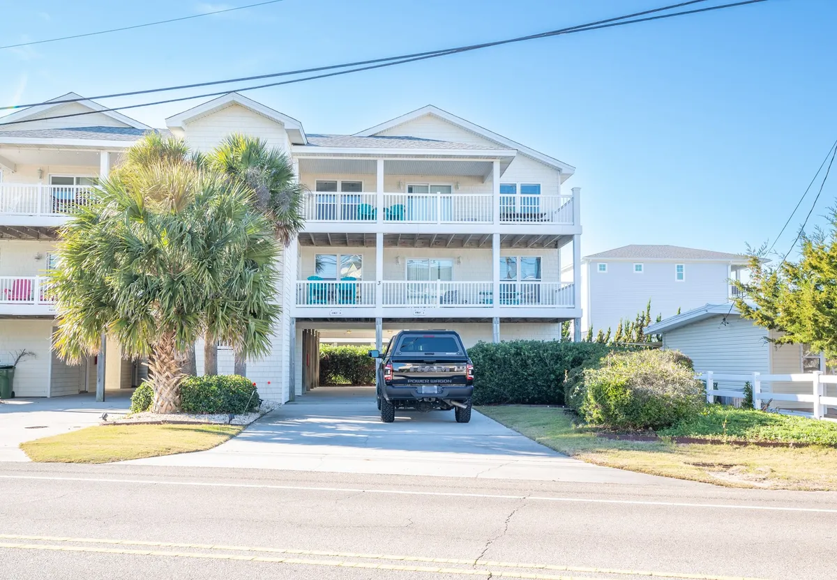 The image showcases the condo’s exterior with balconies furnished with inviting turquoise chairs, a swaying palm tree, and a driveway that hints at easy arrival and beachside living. The scene reflects a relaxed, coastal vibe with outdoor spaces perfect for unwinding while soaking in views and ocean breezes.
