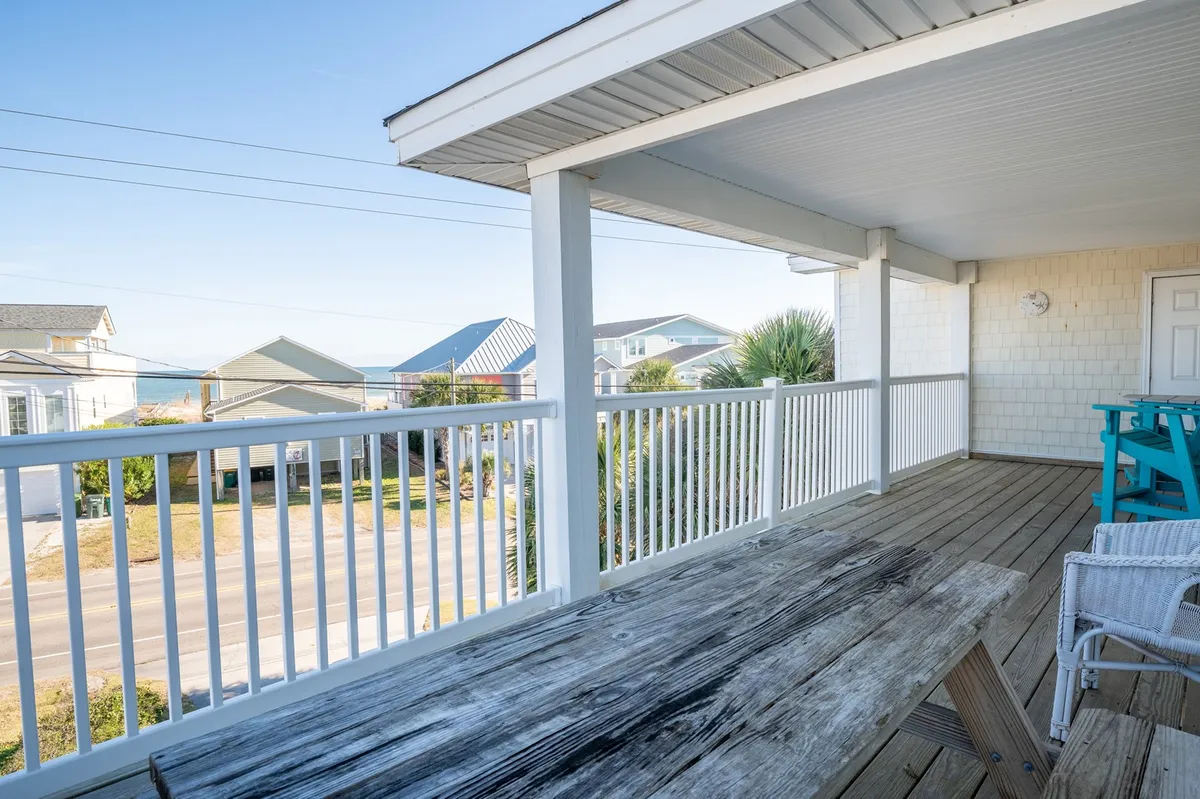 A cozy outdoor dining area featuring a wooden table, blue chairs, and a clear ocean view beyond the white railing. The casual setup is ideal for outdoor meals or unwinding after a day of beach adventures.